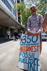 Adam stands at a protest holding a sign that reads "350 Aotearoa/ACC divest/Fossil fuel free"