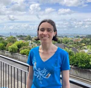 Áine stands smiling outdoors on a house balcony, their short brown hair blowing in the summer breeze. They are a white non-binary femme. Suburban treetops dominate the background, with Auckland's skyline visible in the distance.