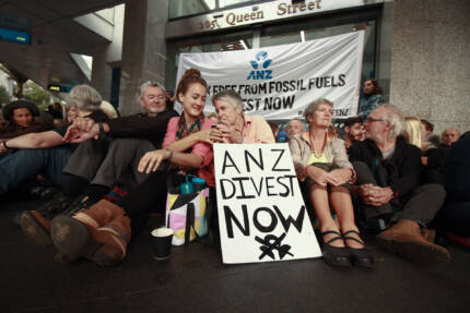Photo of a group of people blocking entrance to ANZ Bank - a sign in the front reads "ANZ divest now" and young women gives an elderly person a hot cup of coffee