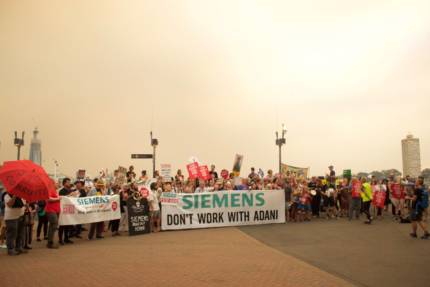 A crowd of 50+ people at a Stop Adani rally calling on Siemens to rule out working with Adani. The sky behind them is hazy with smoke from bushfires