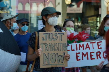 photo of protesters holding signs, person in the front is holding a sign that reads "Just Transition Now"
