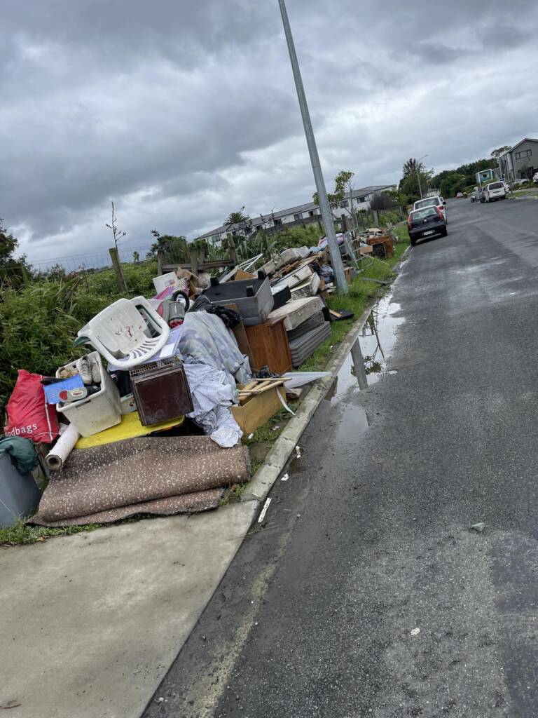 A street on an overcast day is covered in flood damaged furniture and household items on the footpath on the left. 