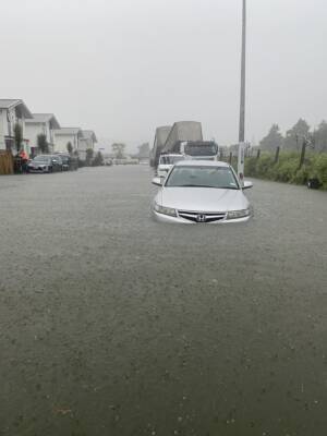 A front on view of a flooded street. Cars are submerged in the water throughout the entire street, the closest car being the deepest (almost up to its headlights). The sky is grey and it continues to rain - water drops are seen on the pool of water from the flood.