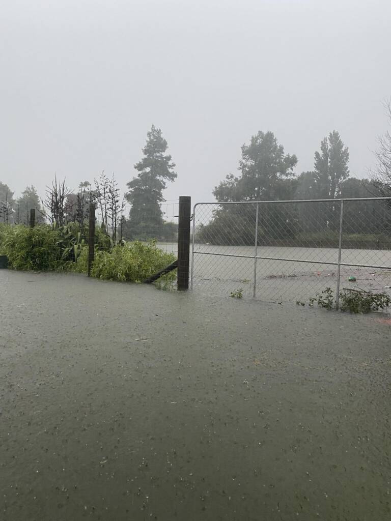 A fence and wired gate stands around severe floods. It is raining on the huge pool of water and the skies are grey.